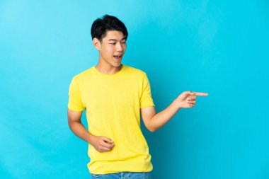 Mavi arka planda izole edilmiş genç bir Çinli parmak göstererek bir ürün sunuyor.