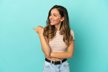 İzole edilmiş mavi arka planda genç bir kadın bir ürünü sunmak için kenarı işaret ediyor.