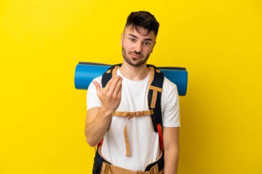 Sarı arka planda izole edilmiş büyük bir sırt çantası olan genç bir Kafkasyalı. Elle gelmeye davet ediyor. Gelmene sevindim.