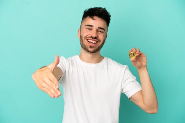 Mavi arka planda izole edilmiş bir Bitcoin tutan genç adam iyi bir anlaşma için el sıkışıyor.