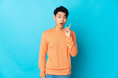 Genç Çinli bir adam mavi arka planda izole edilmiş bir şekilde parmağını kaldırarak çözümü anlamaya çalışıyor.