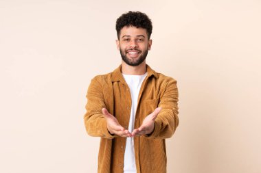 Faslı genç bir adam bej arka planda izole edilmiş. Elinde bir ilan vermek için avuç içi hayali bir şey var.