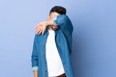 İzole edilmiş mavi arka planda beyaz bir adam elleriyle gözlerini kapatıyor.