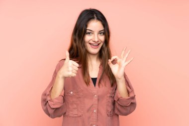 Pembe arka planda izole edilmiş genç beyaz bir kadın, işaret ve baş parmak hareketi gösteriyor.