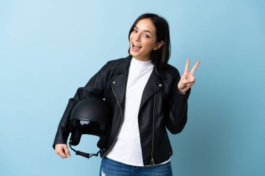 Mavi arka planda izole edilmiş motosiklet kaskı tutan kadın gülümsüyor ve zafer işareti gösteriyor.