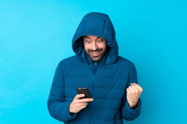 Man wearing winter jacket and holding a takeaway coffee over isolated blue background surprised and sending a message