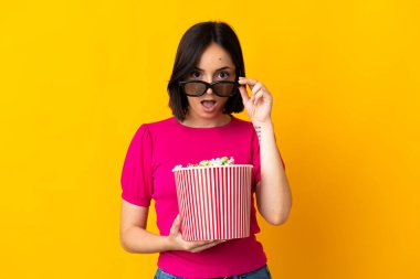 Sarı arka planda izole edilmiş beyaz bir kadın 3D gözlüklü ve elinde koca bir kova patlamış mısırla şaşırdı.