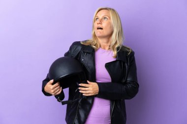 Middle age woman holding a motorcycle helmet isolated on purple background looking up and with surprised expression