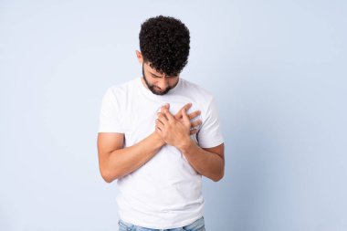 Young Moroccan man isolated on blue background having a pain in the heart