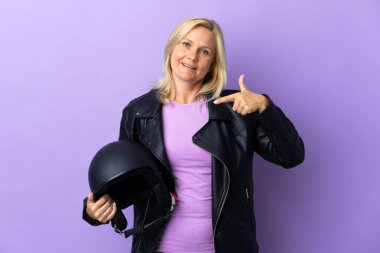 Mor arka planda izole edilmiş motosiklet kaskı tutan orta yaşlı bir kadın baş parmak işareti yapıyor.