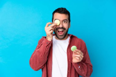 İzole edilmiş mavi arka planda genç beyaz adam renkli Fransız badem kurabiyeleri tutuyor.