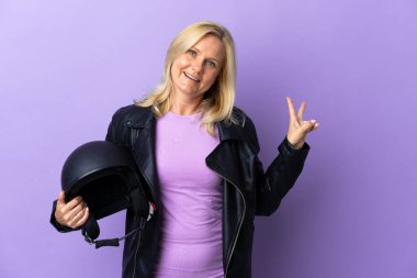 Mor arka planda izole edilmiş motosiklet kaskı tutan orta yaşlı kadın gülümsüyor ve zafer işareti gösteriyor.