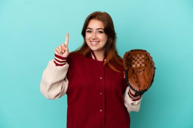 Beyzbol oynayan genç kızıl saçlı kadın mavi arka planda izole edilmiş ve en iyinin işareti olarak parmağını kaldırıyor.