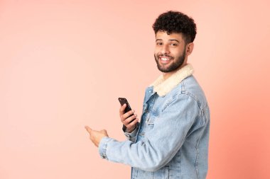 Young Moroccan man using mobile phone isolated on pink background pointing back
