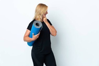 Middle age woman going to yoga classes while holding a mat isolated on white background thinking an idea and looking side