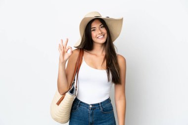 Young Brazilian woman with Pamela holding a beach bag isolated on white background showing ok sign with fingers
