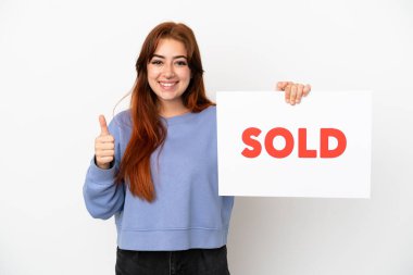 Young redhead woman isolated on white background holding a placard with text SOLD with thumb up