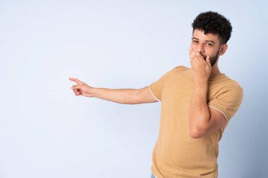 Faslı genç bir adam mavi arka planda izole edilmiş, sürpriz bir ifadeyle işaret ederken...