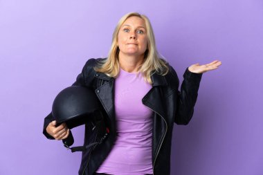 Middle age woman holding a motorcycle helmet isolated on purple background having doubts while raising hands