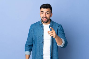 İzole edilmiş mavi arka planı olan beyaz bir adam şaşırmış ve ön tarafı işaret ediyor.