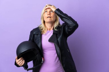 Middle age woman holding a motorcycle helmet isolated on purple background doing surprise gesture while looking to the side