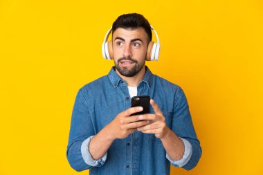İzole edilmiş sarı arka plan üzerinde beyaz bir adam cep telefonuyla müzik dinliyor ve düşünüyor.