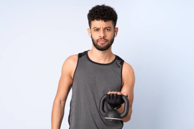 Faslı genç adam mavi arka planda izole edilmiş halter olarak ağırlık kaldırıyor.