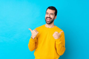 İzole edilmiş mavi bir geçmişi olan genç beyaz bir adam. Baş parmakları havada ve gülümsüyor.