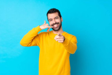 Young caucasian man over isolated blue background making phone gesture and pointing front