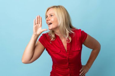 Middle age woman isolated on blue background shouting with mouth wide open to the side
