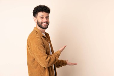 Faslı genç bir adam bej arka planda izole edilmiş. Davet etmek için ellerini yana uzatıyor.