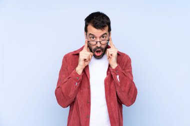 Young caucasian man wearing corduroy jacket over blue background with glasses and surprised