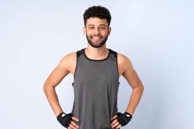 Genç Faslı adam mavi arka planda izole edilmiş. Kolları kalçasında ve gülümsüyor.