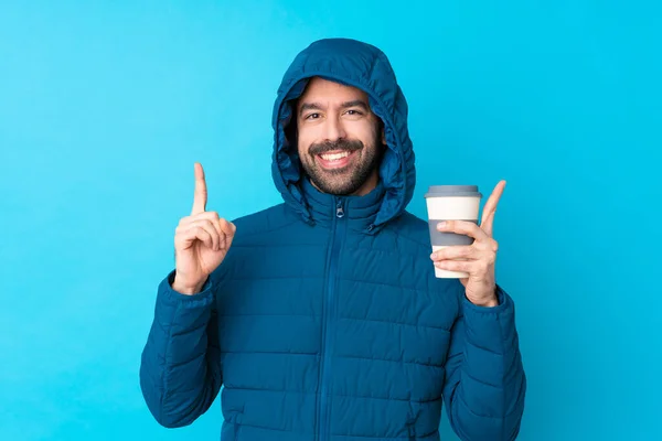 Man wearing winter jacket and holding a takeaway coffee over isolated blue background pointing up a great idea
