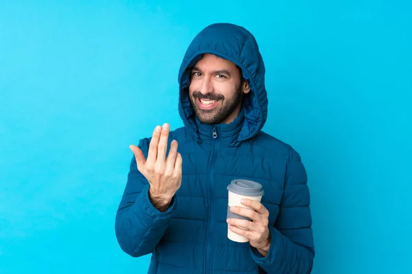 Man wearing winter jacket and holding a takeaway coffee over isolated blue background inviting to come with hand. Happy that you came