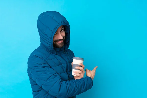 Man wearing winter jacket and holding a takeaway coffee over isolated blue background pointing back
