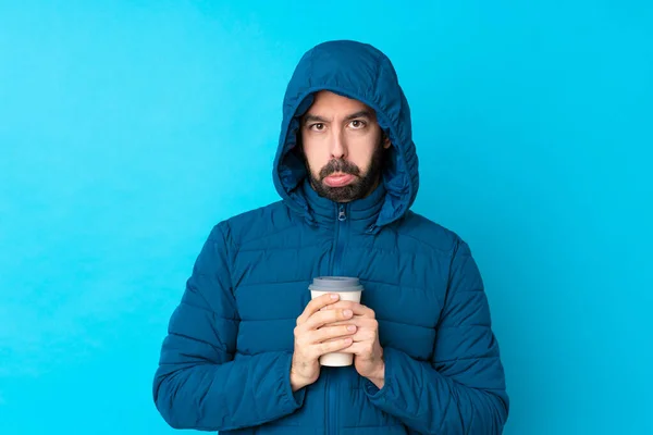 Man wearing winter jacket and holding a takeaway coffee over isolated blue background with sad and depressed expression