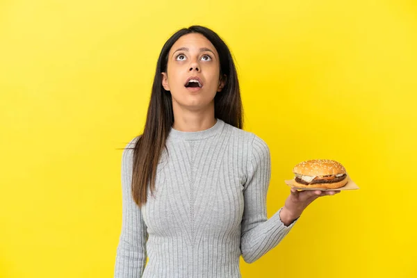 Młoda Kaukaska Kobieta Trzyma Hamburgera Wyizolowany Żółtym Tle Patrząc Górę — Zdjęcie stockowe