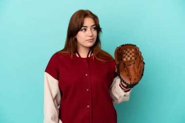 Young redhead woman playing baseball isolated on blue background looking to the side
