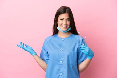 Pembe arka planda izole edilmiş aletleri tutan dişçi kadın davet edilmek için ellerini yana uzatıyor.