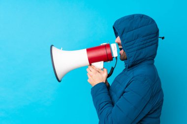 Kışlık ceket giyen ve elinde kahve tutan bir adam izole edilmiş mavi arka planda megafonla bağırıyor.
