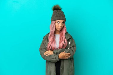Pembe saçlı, yağmura dayanıklı bir ceket giyen, mavi arka planda izole edilmiş, kollarını kavuşturan genç bir kadın.