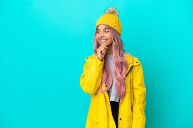 Pembe saçlı, yağmura dayanıklı bir ceket giyen genç bir kadın mavi arka planda izole edilmiş bir şekilde yan tarafa bakıyor ve gülümsüyor.