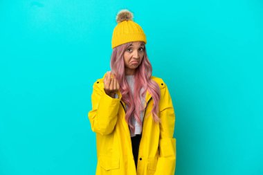 Pembe saçlı, yağmura dayanıklı bir ceket giyen, mavi arka planda izole genç bir kadın para hareketi yapıyor.