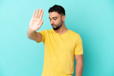 Genç Arap adam mavi arka planda izole edilmiş. Durma hareketi yapıyor ve hayal kırıklığına uğruyor.