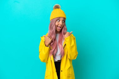 Pembe saçlı, yağmura dayanıklı bir ceket giyen genç bir kadın. Mavi arka planda izole edilmiş. Baş parmakları havada ve gülümsüyor.