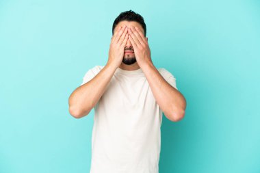 Genç Arap adam mavi arka planda izole edildi. Gözlerini elleriyle kapadı.