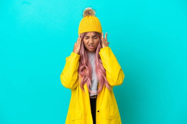 Pembe saçlı, yağmura dayanıklı bir ceket giyen, mavi arka planda başağrısı olan genç bir kadın.