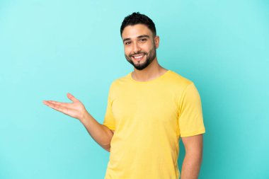 Genç Arap adam mavi arka planda izole edilmiş bir şekilde gülümserken bir fikir sunuyor.