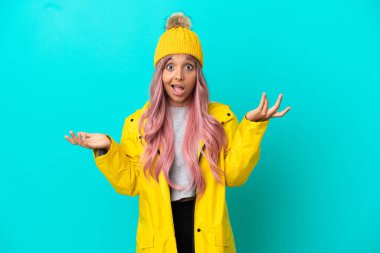 Pembe saçlı, yağmura dayanıklı bir ceket giyen genç bir kadın. Mavi arka planda izole edilmiş, şok olmuş bir yüz ifadesi var.